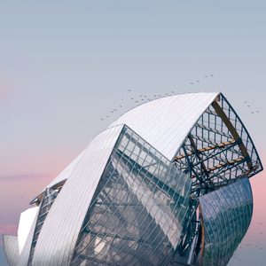 Photographie de la Fondation Louis Vuitton avec sa structure moderne en verre et métal, capturée au coucher du soleil avec des teintes douces de rose et bleu dans le ciel. Le design courbé et géométrique du bâtiment est mis en valeur, soulignant la complexité et l'élégance de son architecture. Un vol d'oiseaux traverse le ciel, ajoutant du mouvement à cette scène sereine.