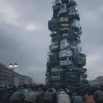 Totem fait de voitures empilées avec des personnes en prière, symbolisant le consumérisme et l'adoration des objets matériels