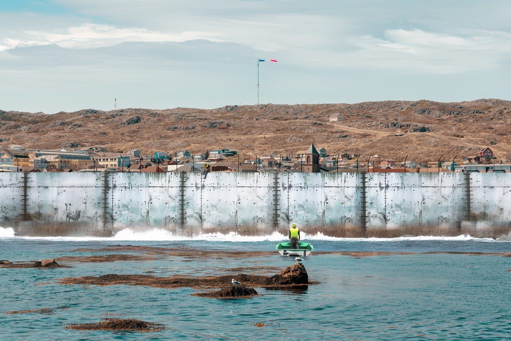 View of a walled city on a rocky landscape, with a person in a small boat near the water's edge, and a barbed-wire fence on top of the weathered concrete wall.