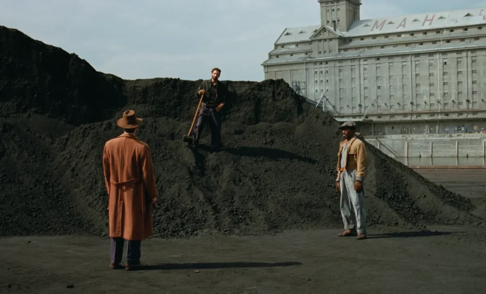 Trois hommes se font face dans un paysage industriel poussiéreux, entourés de monticules de charbon. L’un d’eux, en manteau long et chapeau, tourne le dos à la caméra, tandis qu’un autre, en tenue de travail, est juché sur un tas de charbon, tenant un marteau. En arrière-plan, un immense bâtiment brutaliste aux allures de silo domine la scène, portant des lettres partiellement visibles sur son toit.