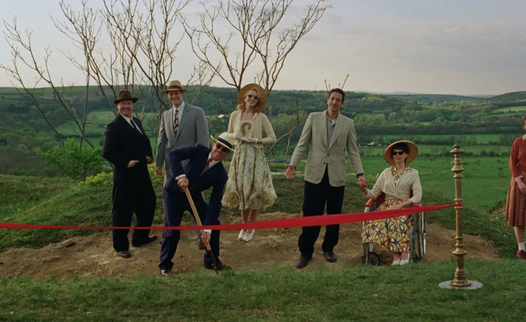 Un groupe de six personnes vêtues de tenues élégantes des années 1950 participe à une cérémonie de pose de première pierre sur une colline verdoyante. Un homme en costume sombre et chapeau creuse avec une pelle, tandis qu’une femme en fauteuil roulant et d’autres convives sourient. L’arrière-plan révèle une campagne vallonnée sous un ciel légèrement nuageux.