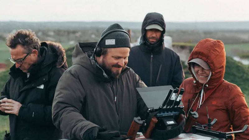 Une équipe de tournage, habillée chaudement, travaille en extérieur sur un plateau de The Brutalist. Au centre, un réalisateur souriant consulte un écran de contrôle, entouré de techniciens et d’une cadreuse équipée d’un rig de stabilisation. L’arrière-plan montre un paysage rural sous un ciel couvert.